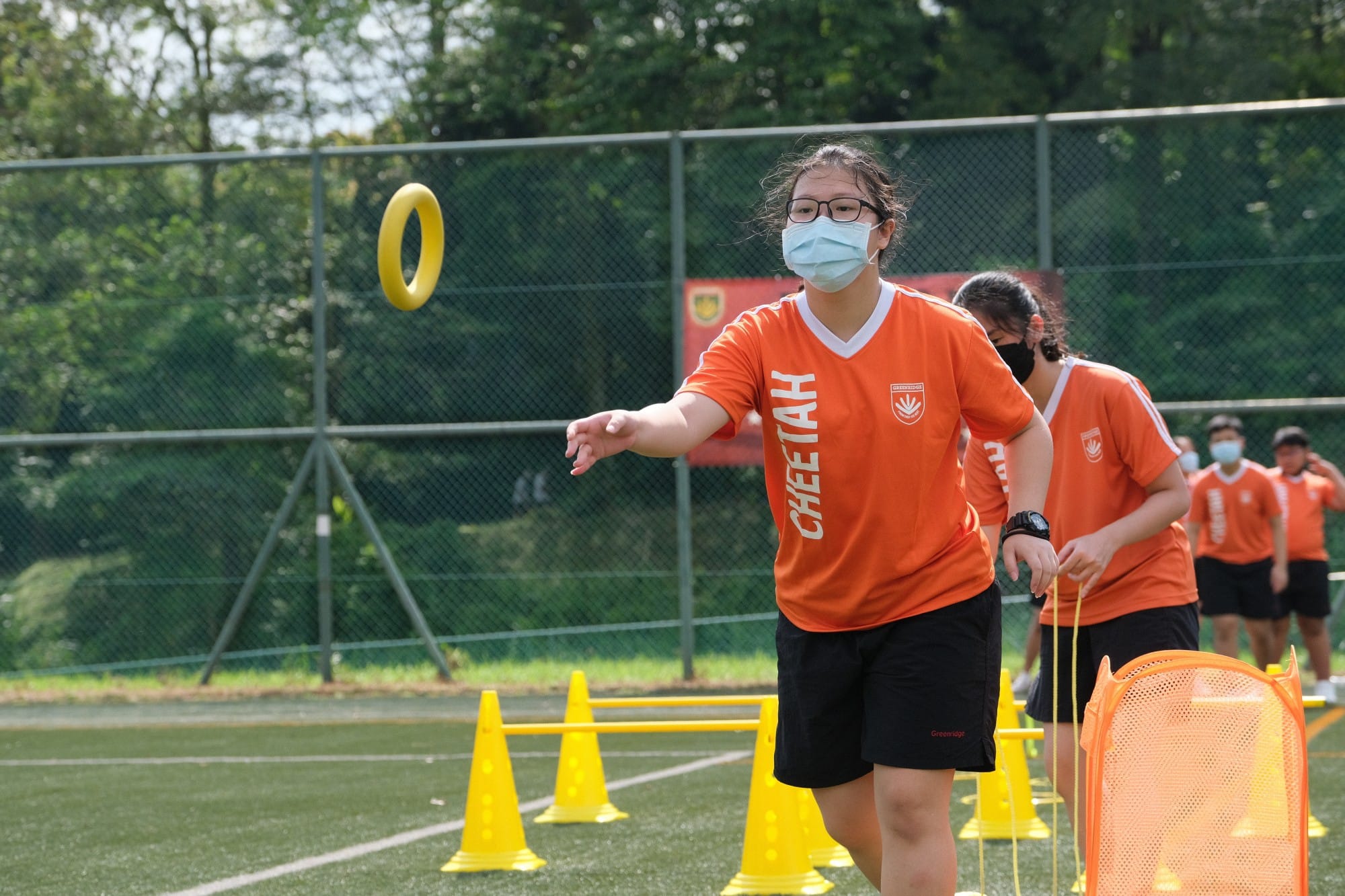 A masked girl in an orange Cheetah jersey throws a yellow ring toward a target on an outdoor sports field.